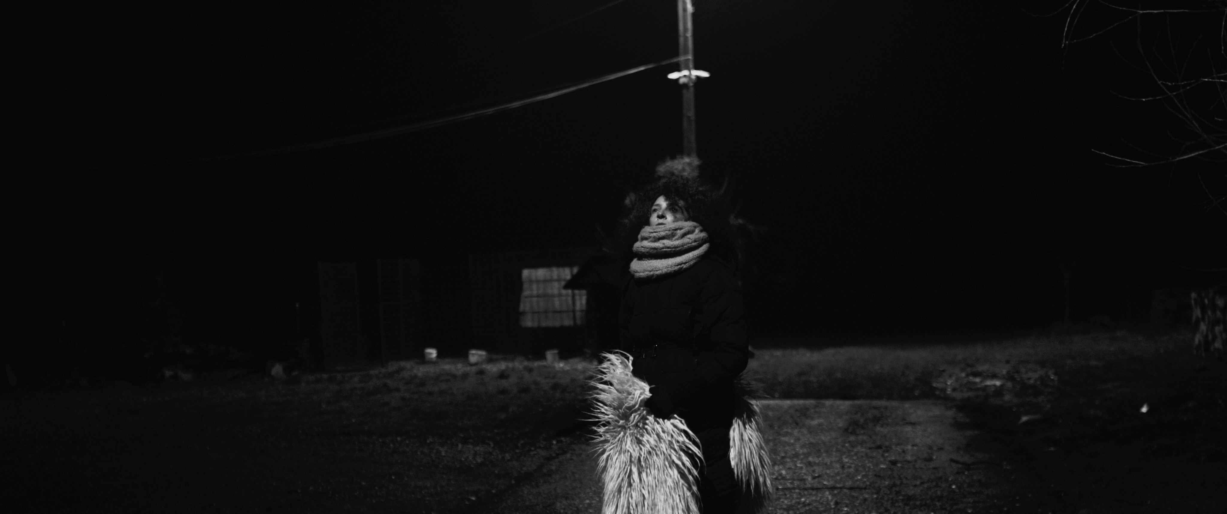 Black and white photograph of Gheorghe in the foreground on the right-hand side of the picture playing the violin on a bench. He is wearing a white t-shirt with black braces. A guitar is drawn on his braces. He is in a park with fallen leaves on the grass. In the background, a street with traffic and communist buildings.