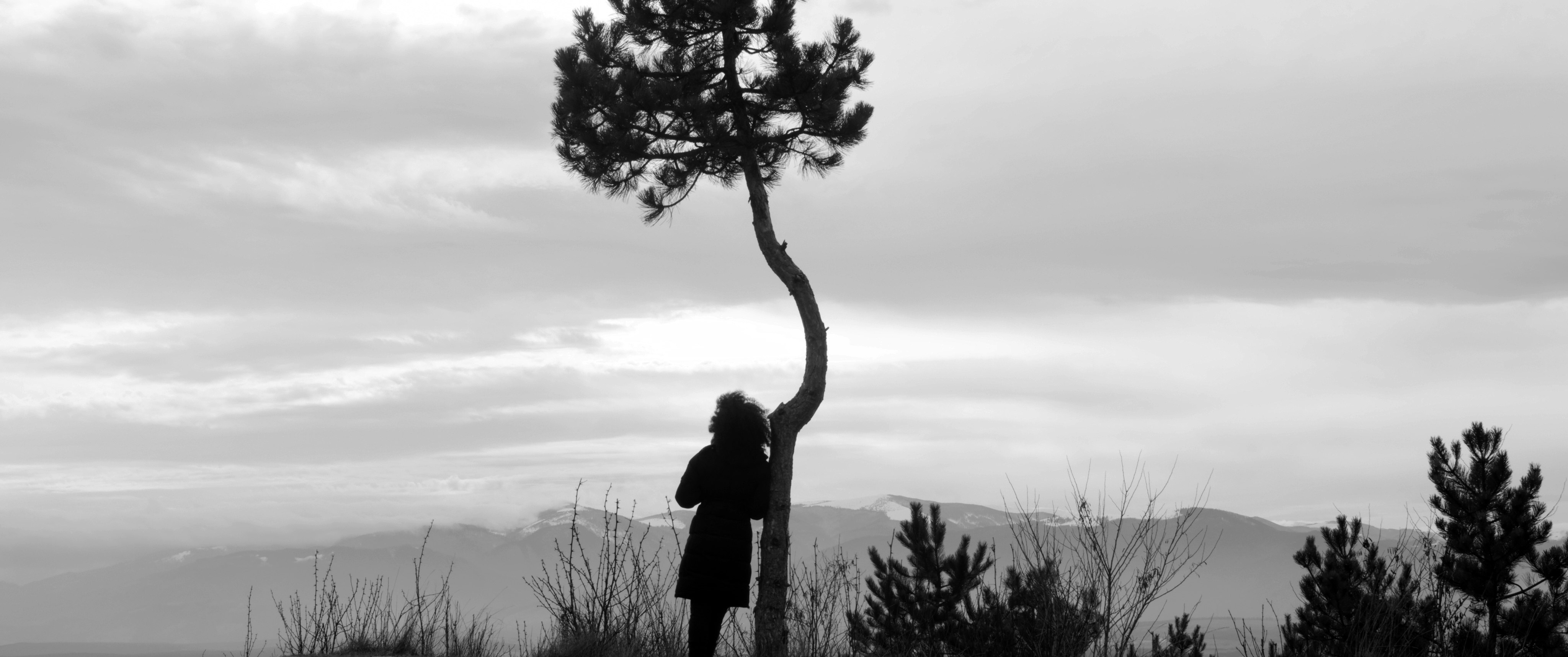 Black and white photograph of Gheorghe sitting at a table in the courtyard of his house. He is looking at the camera, talking and smiling. His left arm is resting on the table, his right hand is raised.