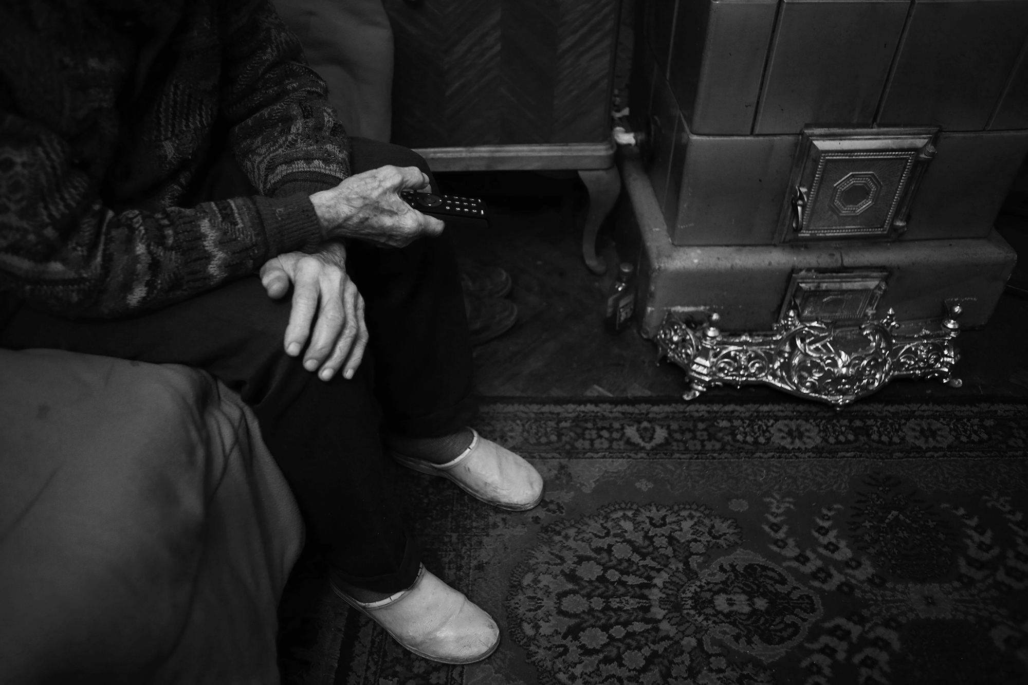 Black and white photograph of Licu on the left, sitting on a bed. His face is not visible. His hands are crossed. The right hand is holding a remote control, the left is resting on the right knee.