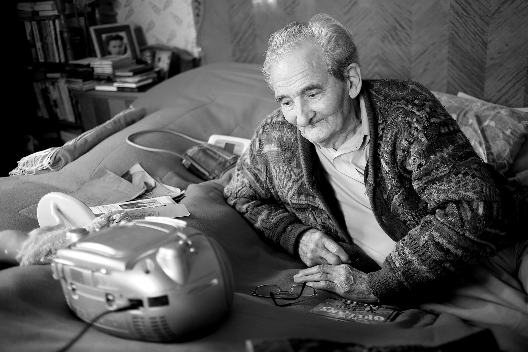 Black and white photograph of Licu leaning on his bed with his right arm. He is watching a radio-CD. Albums lie under his right hand. In the background, a photograph of his wife is on a bedside table.