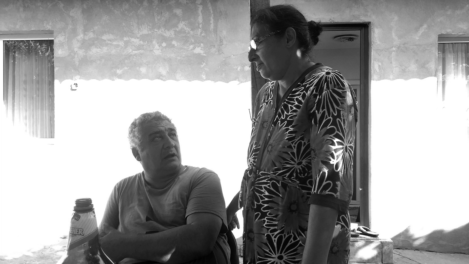 Black and white photograph of Gheorghe and Sorina, his wife. Gheroghe is sitting at a table in the courtyard of their house, talking and looking at her. Sorina is standing with her hand on the back of the chair, looking at him.