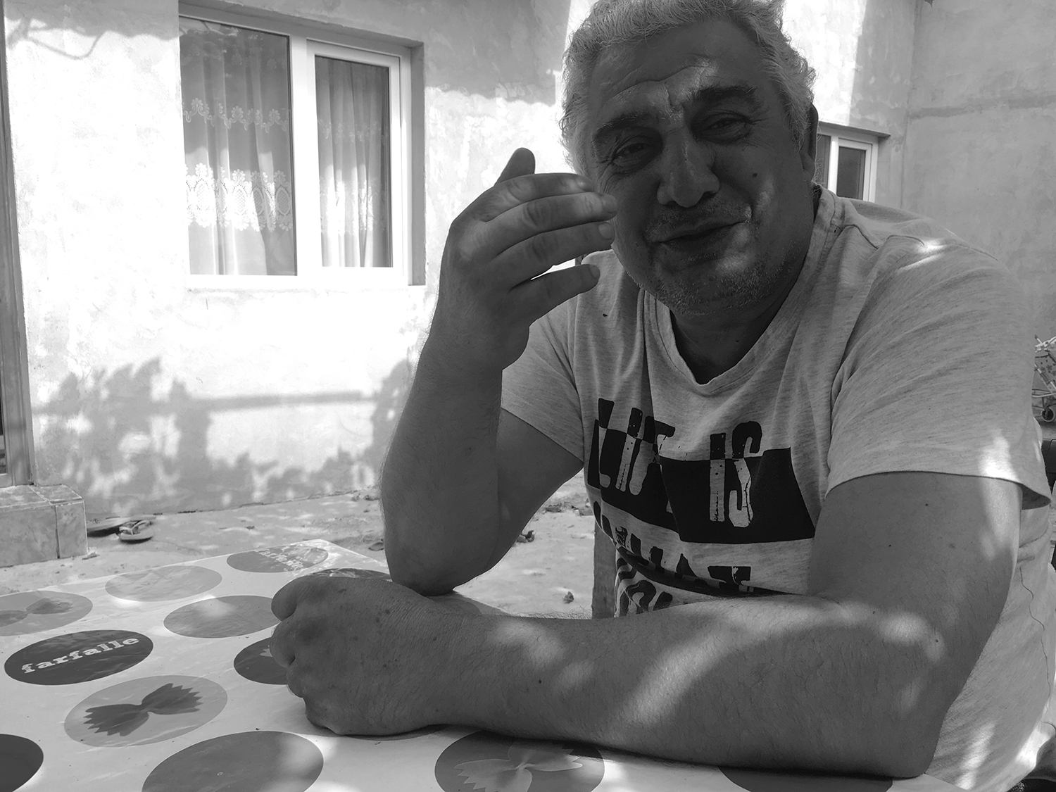 Black and white photograph of Gheorghe sitting at a table in the courtyard of his house. He is looking at the camera, talking and smiling. His left arm is resting on the table, his right hand is raised.