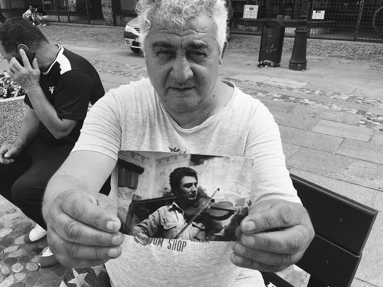 Black and white photograph of Gheorghe sitting on a bench. He is staring into the camera and holding out a photograph of himself playing the violin when he was younger. 
